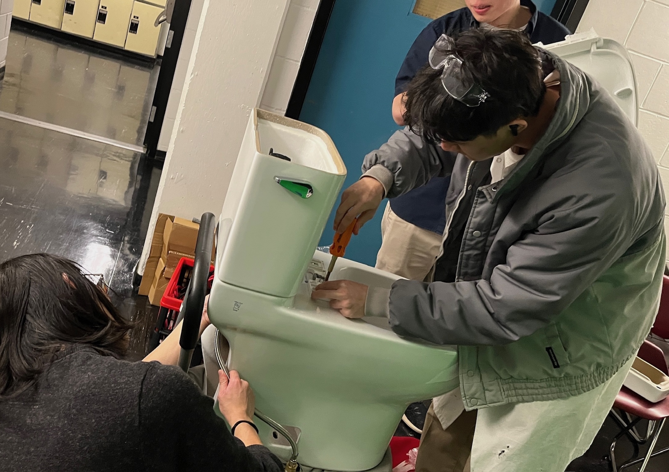 The team is assembling the first build of Orbital Flush in a random hallway. Kai is screwing in the toilet tank hose, while Bill and Calvin affix the toilet seat with a screwdriver.