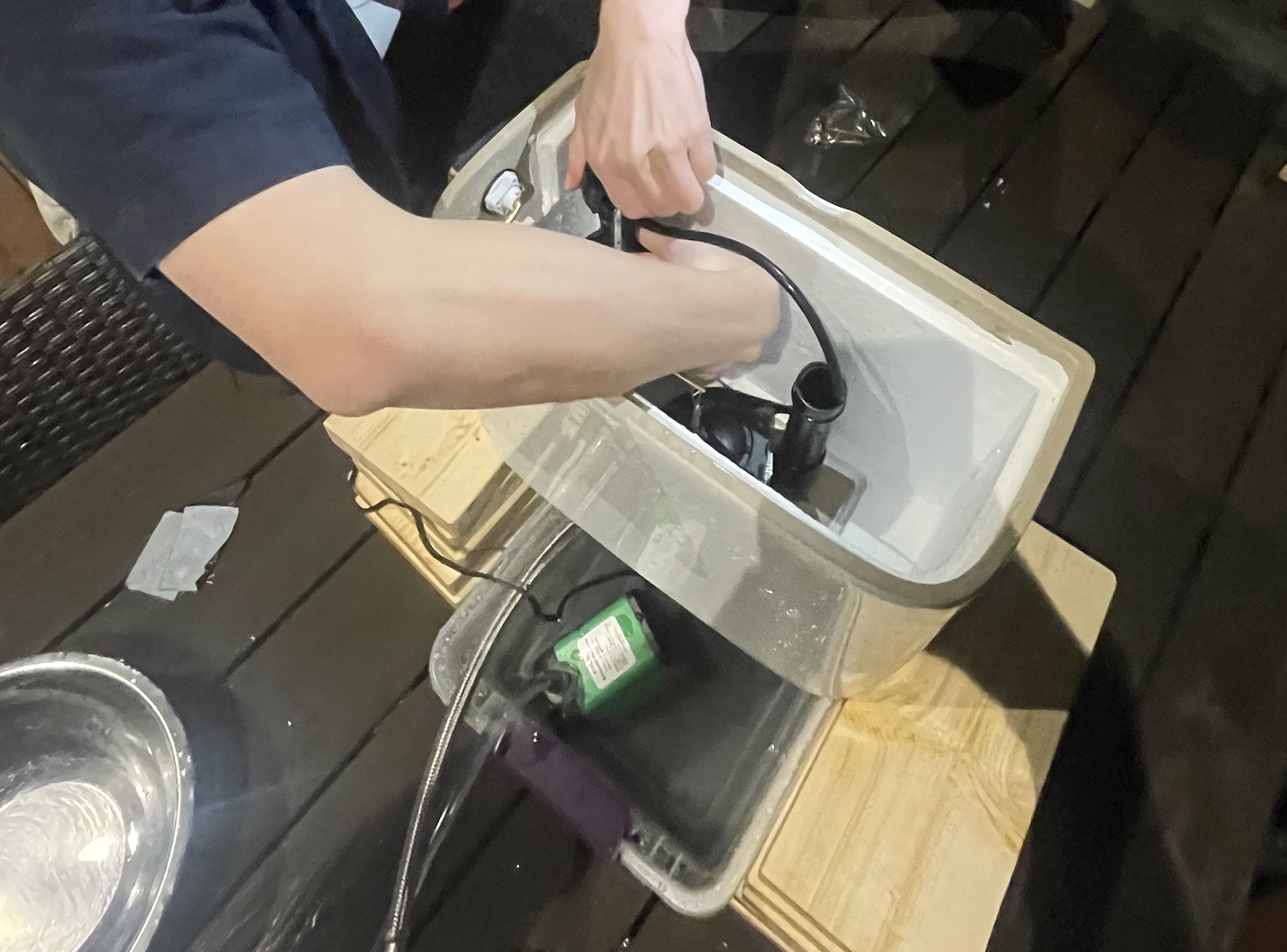 Top-down view of a toilet tank propped up on top of wooden planks atop a plastic container. A small water pump is inside the plastic container, connected to the toilet fill valve.
